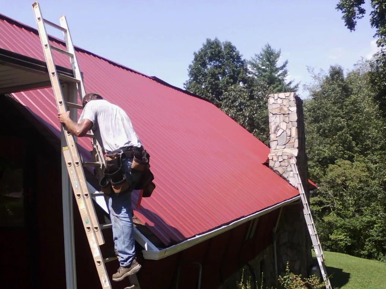 Roofer on a ladder beside a red metal roof and stone chimney — Soffit & Fascia Repair by Roofing Brothers in Bayside